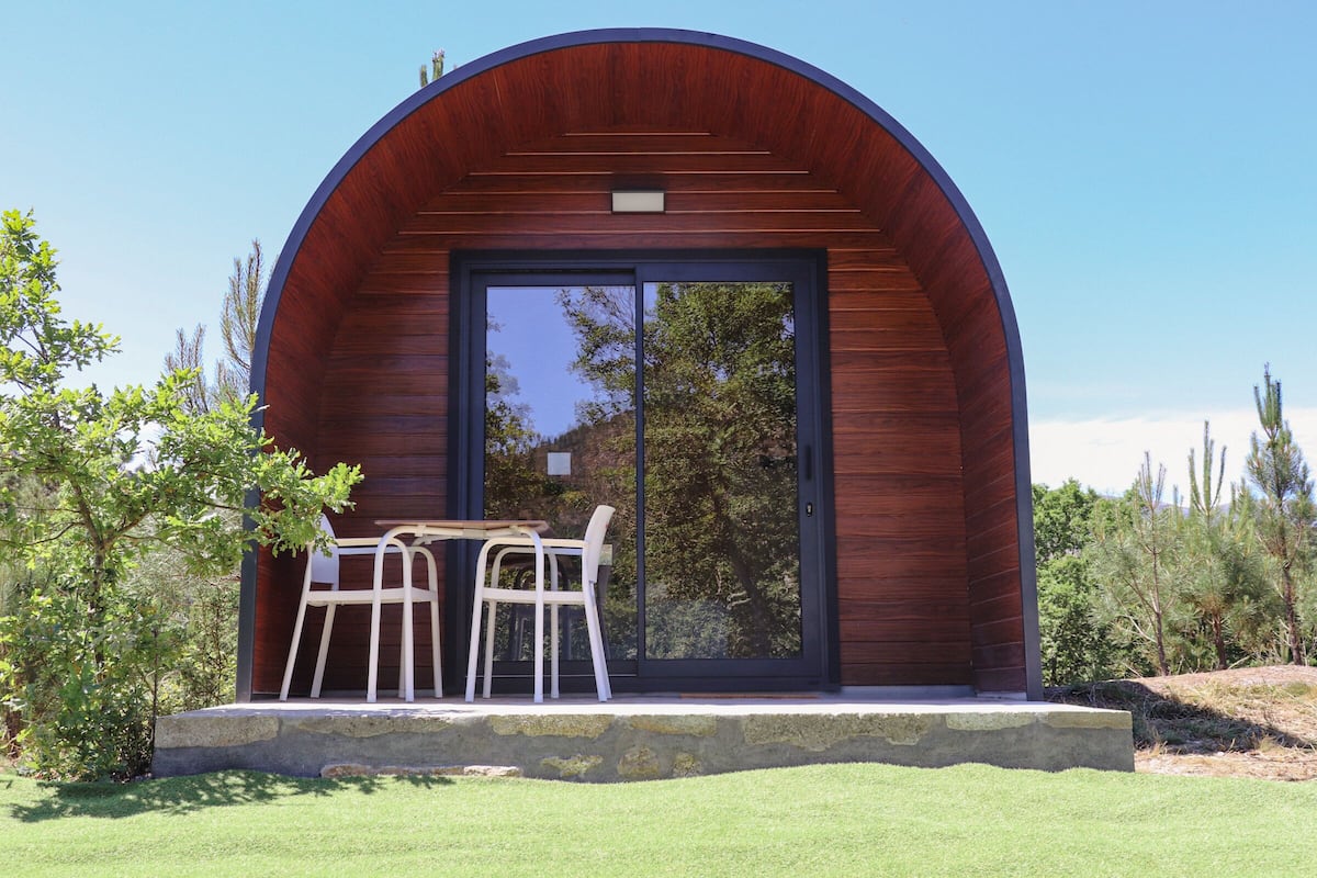 A modern building with a curved wooden facade and a glass door. There is a small outdoor seating area with two chairs and a table.