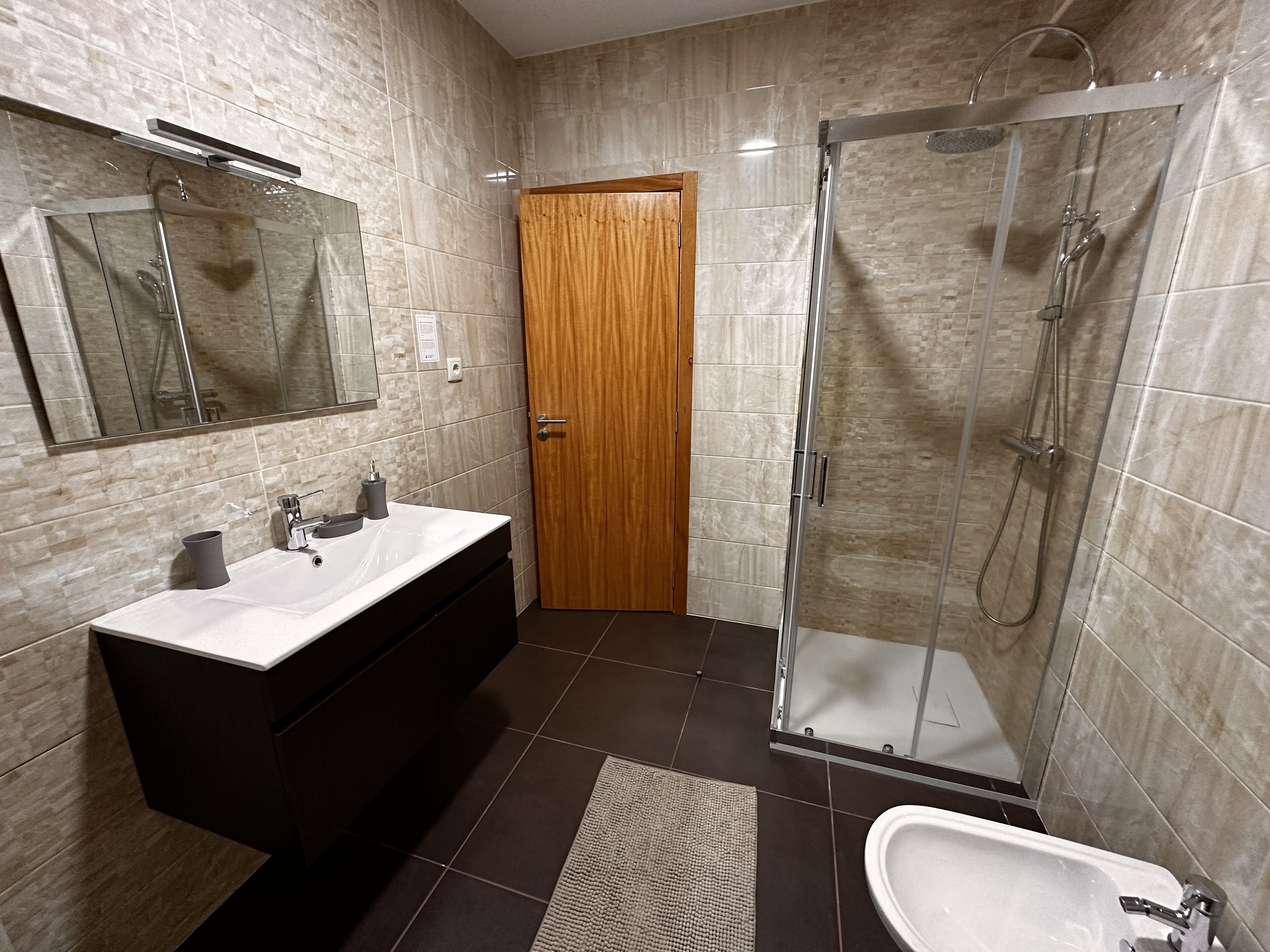 A modern bathroom with a glass-enclosed shower, a white sink with a dark cabinet, and a wooden door.