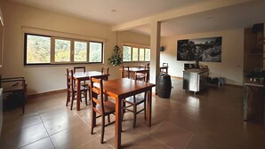 Un vaste espace dédié à la salle à manger, avec une grande table et des chaises en bois, une plante suspendue et une vue sur les arbres par les fenêtres.