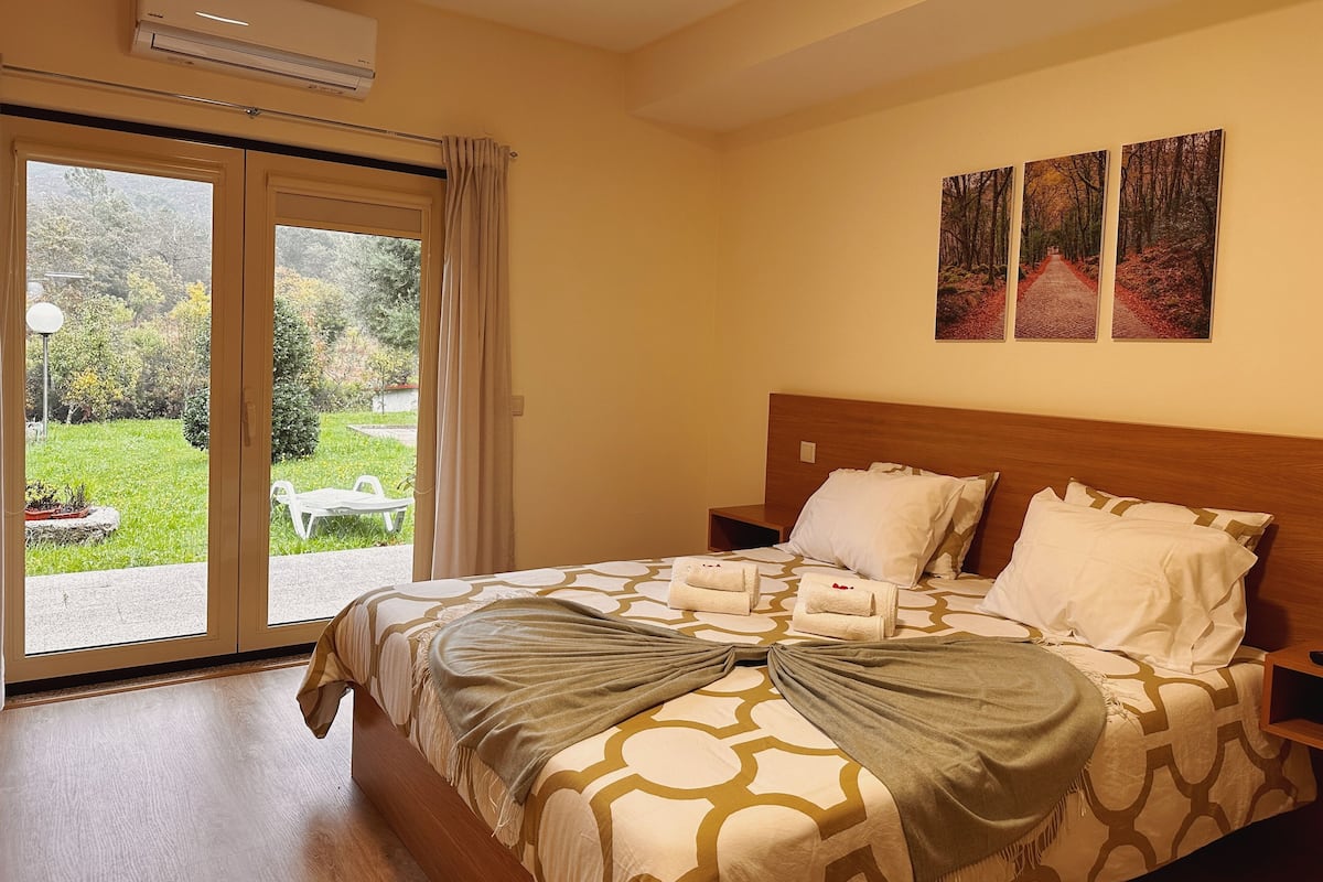 A hotel room with a bed, a nightstand, a window with curtains, and a view of a garden.