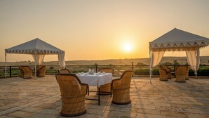 Une vue sur le coucher de soleil depuis une terrasse où une table est dressée pour deux, entourée de chaises en osier, sous une tonnelle.