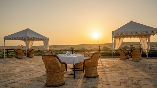 Een zonsondergang uitzicht vanaf een terras met een gedekte tafel voor twee, omringd door rieten stoelen, onder een overkapping.