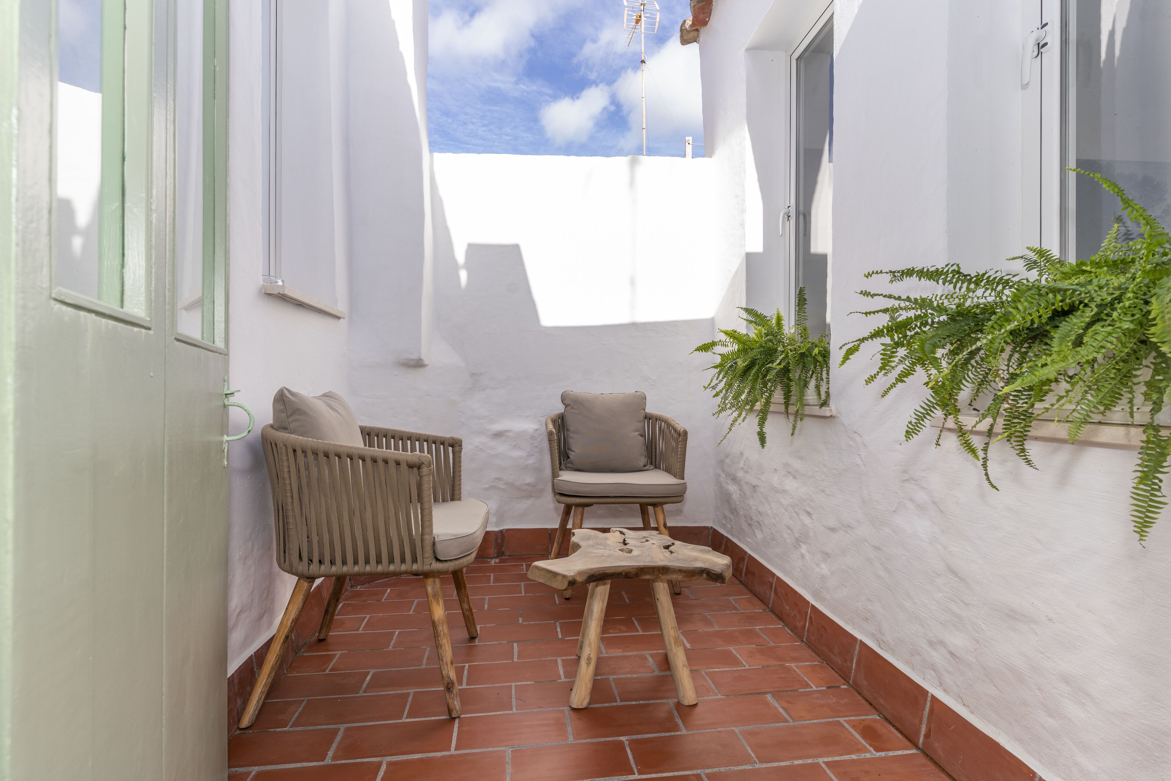 Un pequeño patio con dos sillas de mimbre y una mesa de madera, paredes blancas y piso de baldosas.