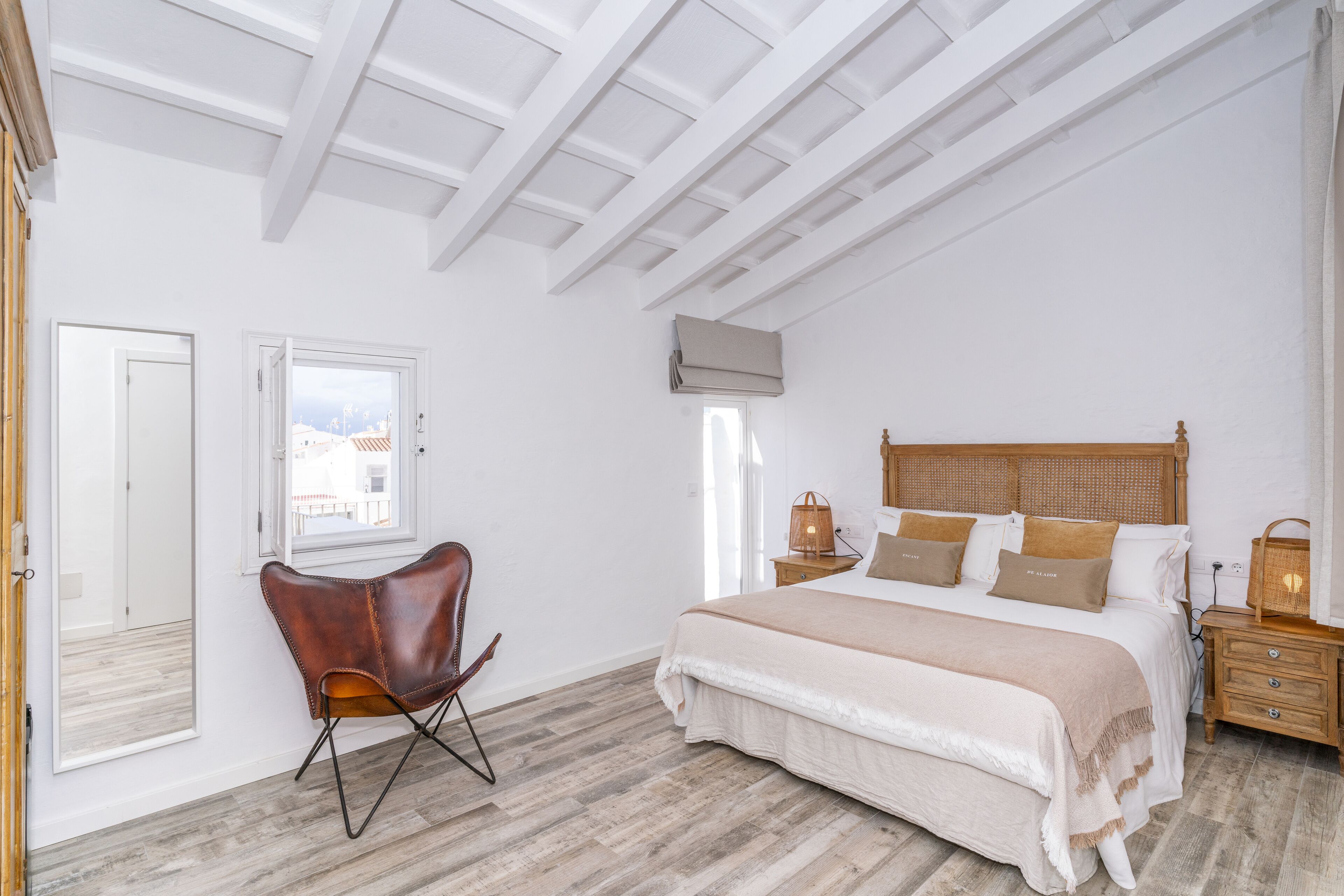 Un dormitorio con una cama grande, una mesita de noche de madera, una silla y una ventana con vistas a edificios.