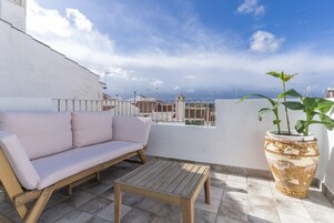 Un balcón con pared blanca, sofá color arena, mesa de madera y una planta grande en maceta.