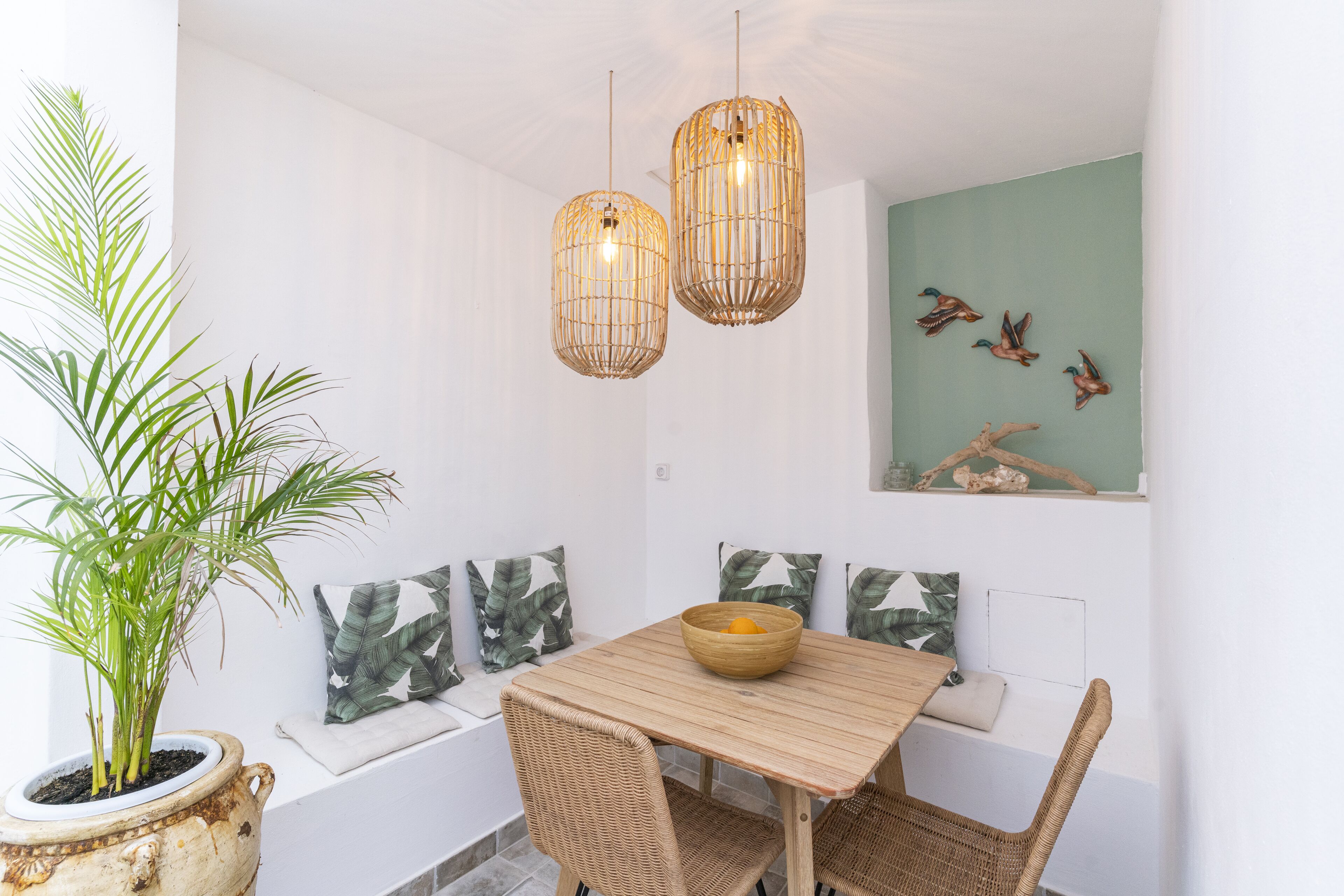 Un comedor con mesa de madera, sillas de mimbre, una planta en maceta y lámparas colgantes de mimbre.
