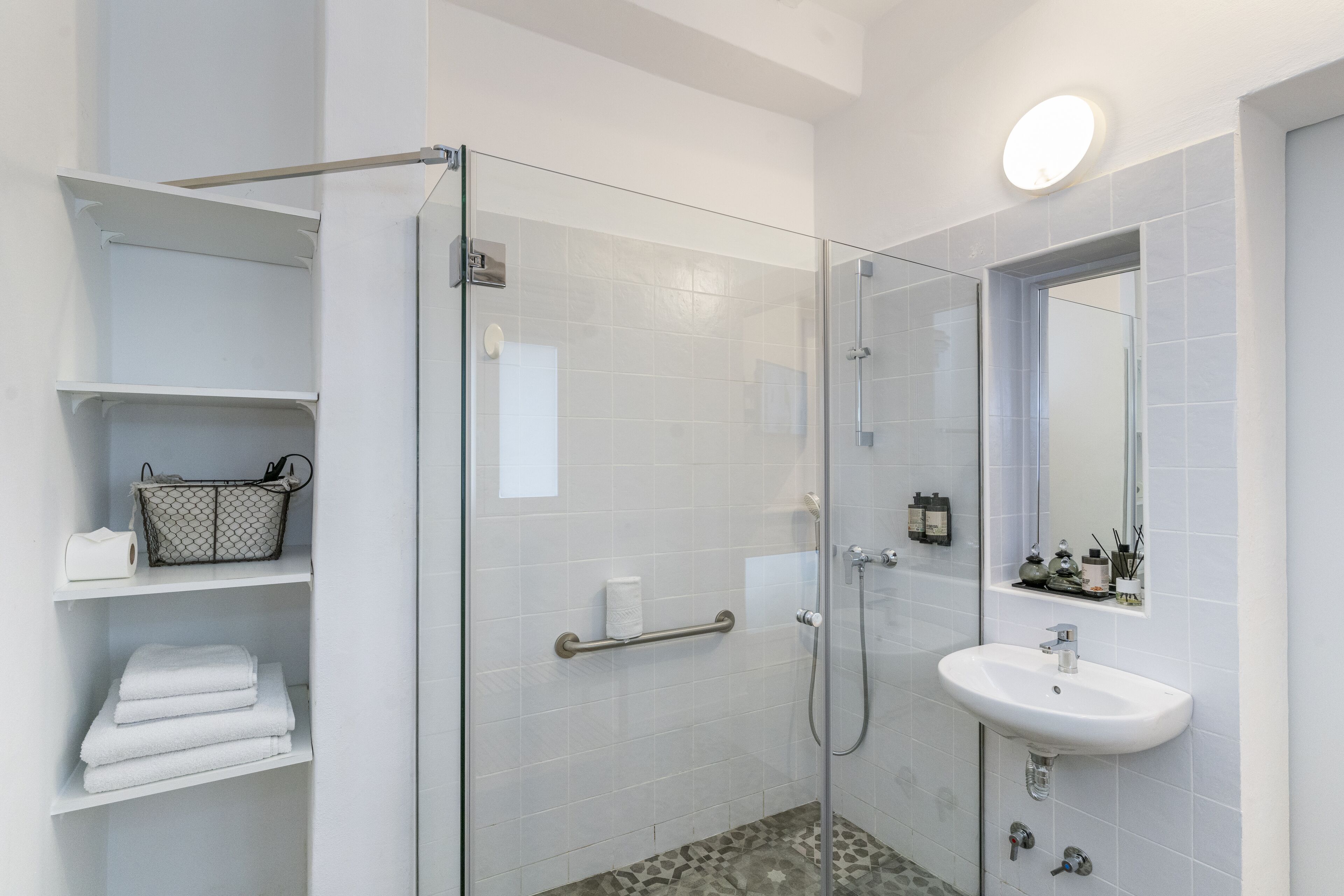 A modern bathroom with a glass shower enclosure, white tiled walls, a white sink, and a mirror above it. There are shelves with towels and toiletries.