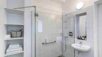 A modern bathroom with a glass shower enclosure, white tiled walls, a white sink, and a mirror above it. There are shelves with towels and toiletries.