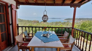 Une table en bois recouverte d’une nappe bleue, entourée de chaises, sur un balcon offrant une vue sur la mer et les collines.