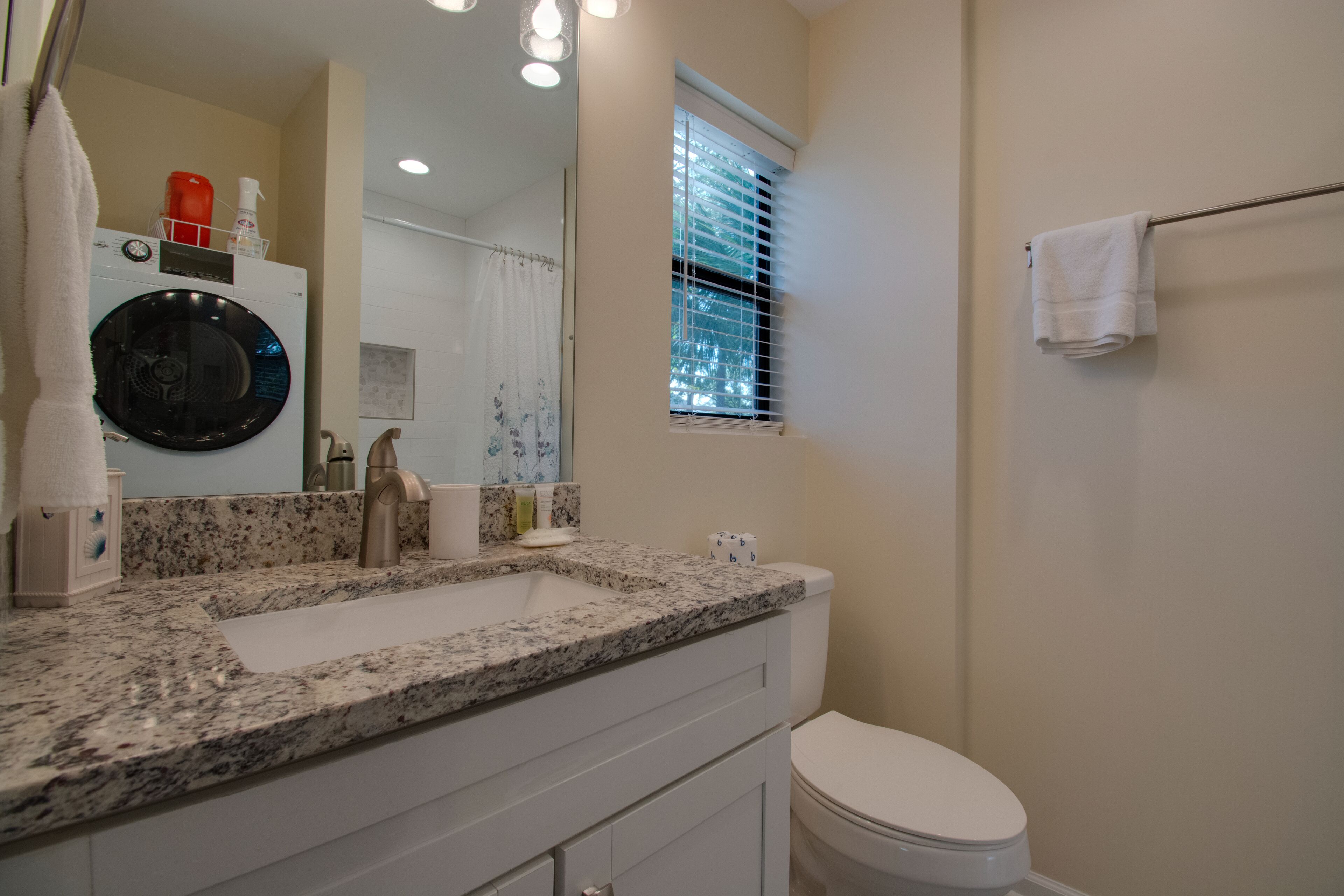 A bathroom with a granite countertop sink, a toilet, a towel rack, and a window with blinds.