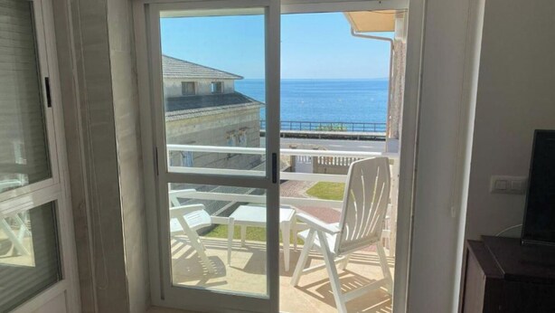 A room with a sliding glass door leading to a balcony with white chairs and a view of the sea. - LAUNCHED HOLIDAYS (Sanxenxo)