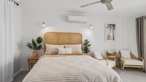 A bedroom with a checkered bedspread, a wicker headboard, bedside tables with plants, a ceiling fan, an air conditioning unit, and a wall-mounted artwork.