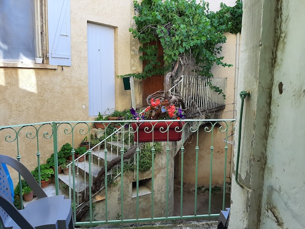 Une cour étroite, fermée par un portail en métal, agrémentée de plantes en pot et dotée d’un escalier menant à un bâtiment avec une porte blanche et des volets aux fenêtres.