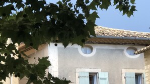 Un bâtiment avec un toit en tuiles, deux fenêtres avec volets et un arbre dont le feuillage obstrue partiellement la vue.
