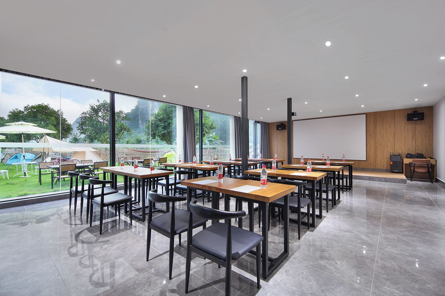 A modern conference room with tables and chairs, large windows, and a projector screen.