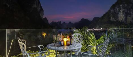 A balcony with a table set for two, surrounded by white metal chairs, overlooking a mountainous landscape at dusk.