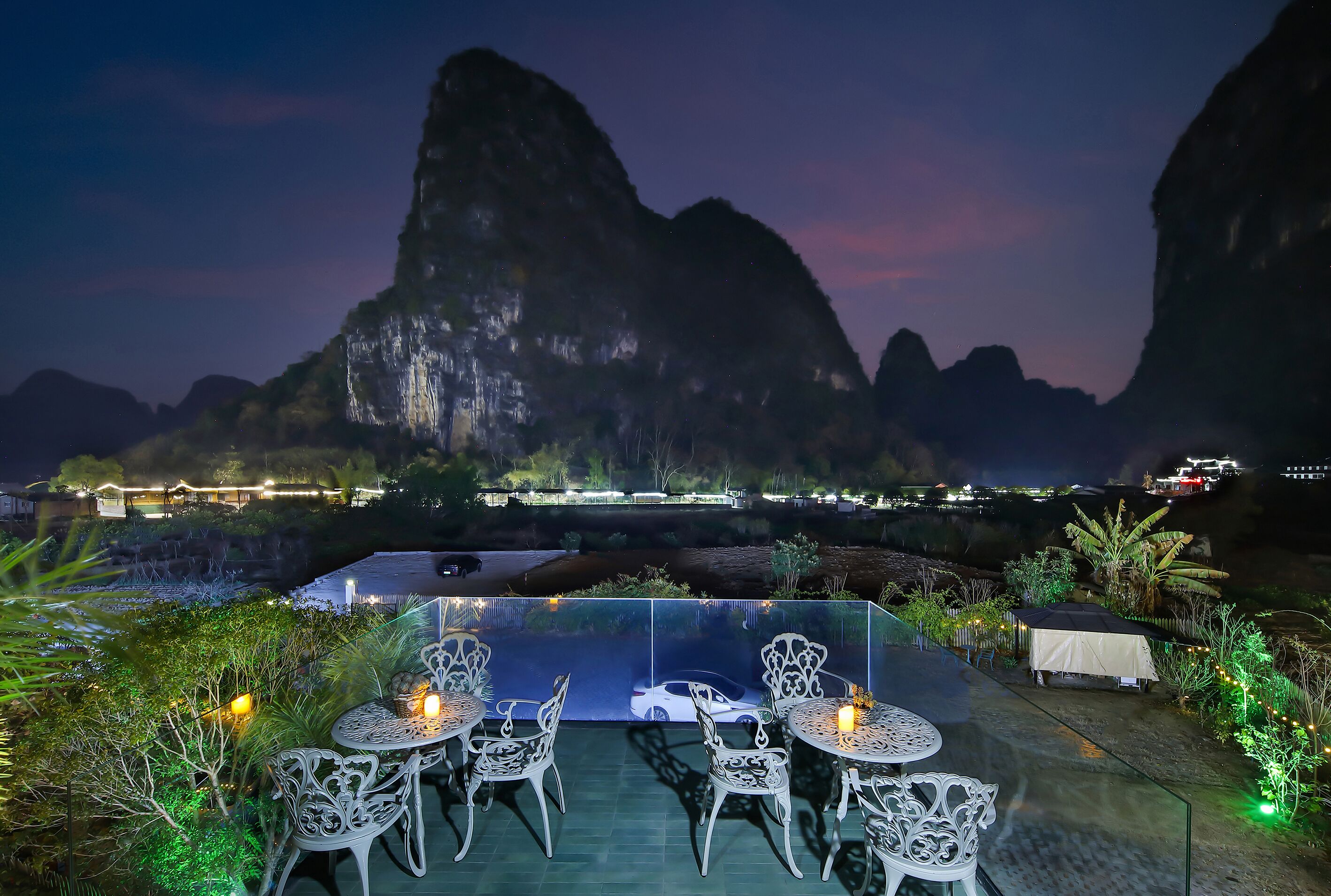 Una terraza en la azotea con barandilla de vidrio, sillas blancas y una mesa, con vistas a un paisaje montañoso al atardecer.
