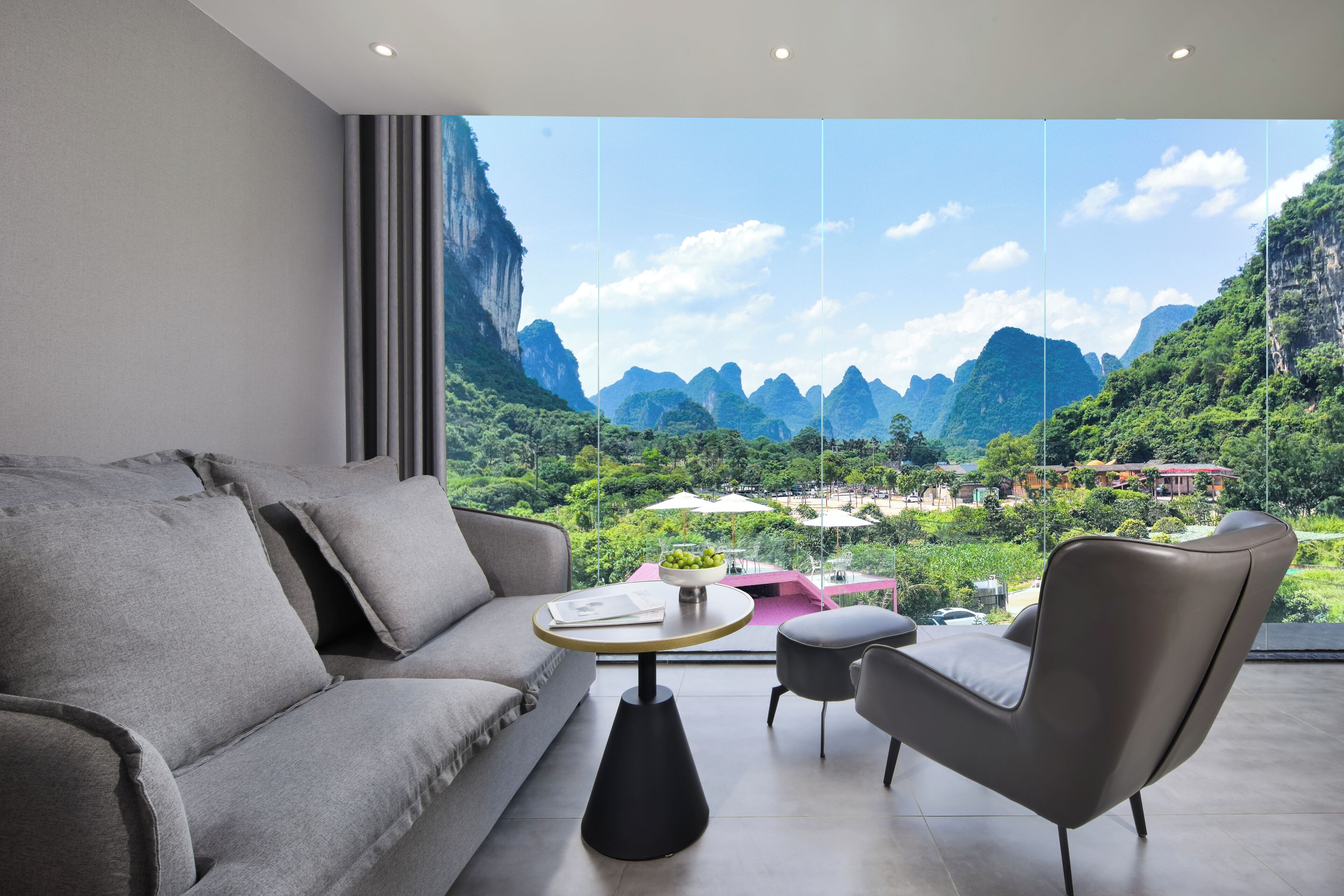 A modern room with a sofa, a small round table, and a chair, with a large window offering a view of mountains and greenery.