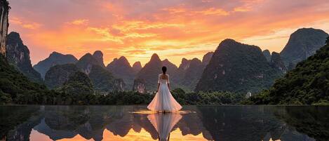 A woman in a white dress standing by a reflective lake with mountainous scenery in the background.