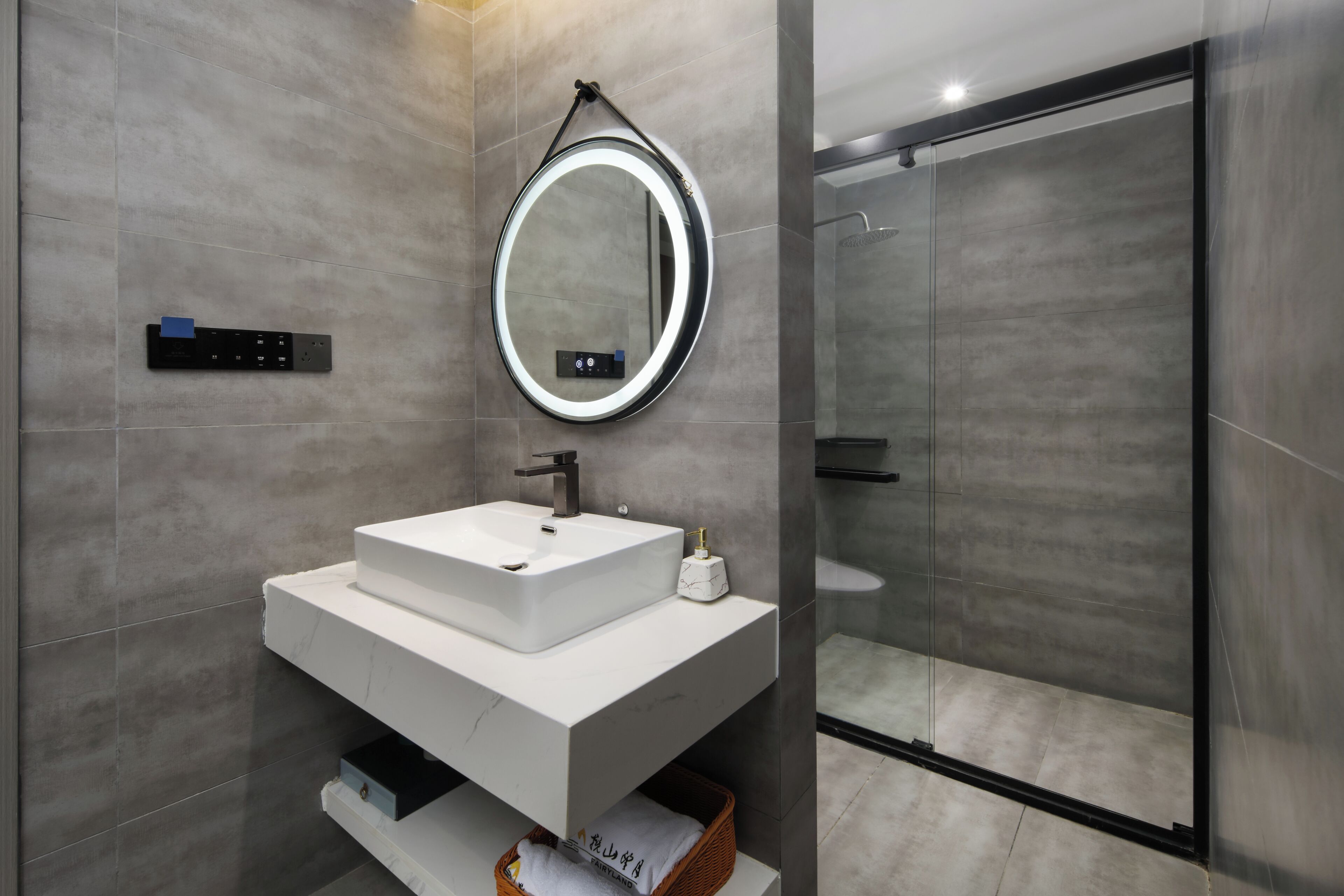 A modern bathroom with a white sink, a round mirror with a light, and a glass shower enclosure.