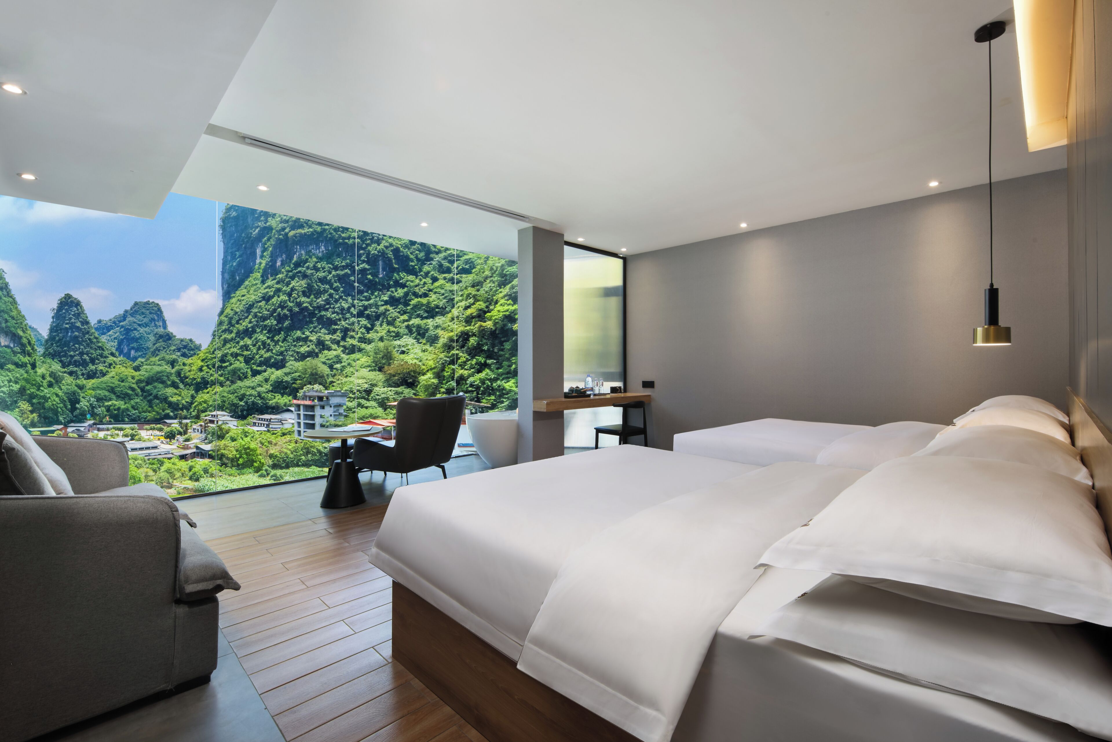 A modern hotel room with a large bed, a desk, a chair, and a view of a lush green landscape.