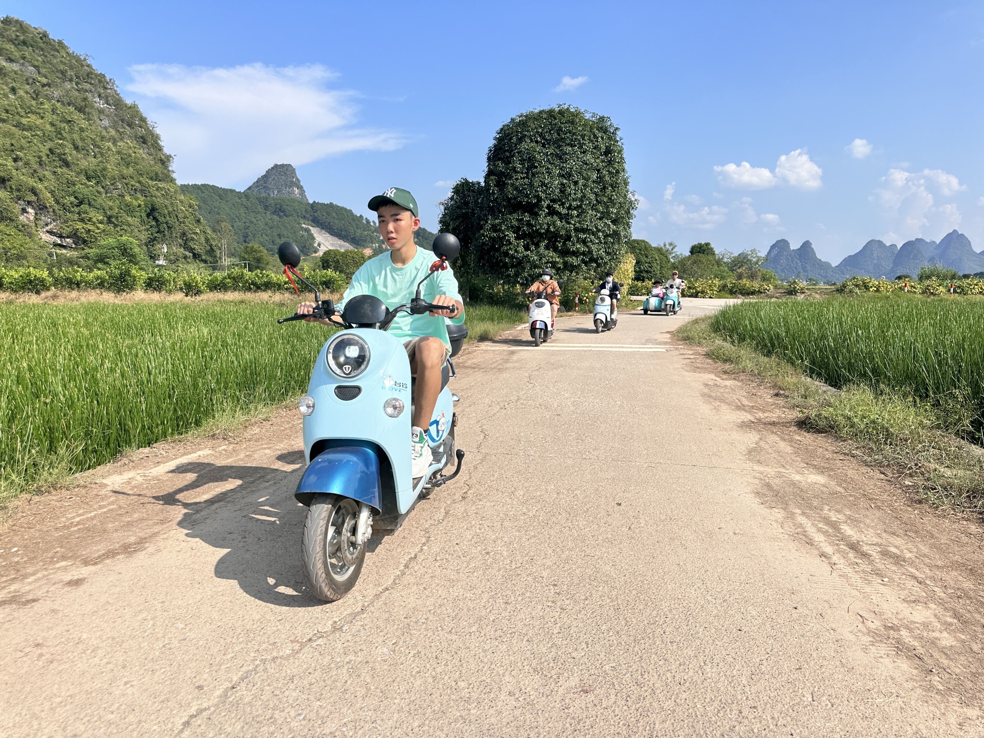 A person riding a scooter on a dirt road with fields and mountains in the background.