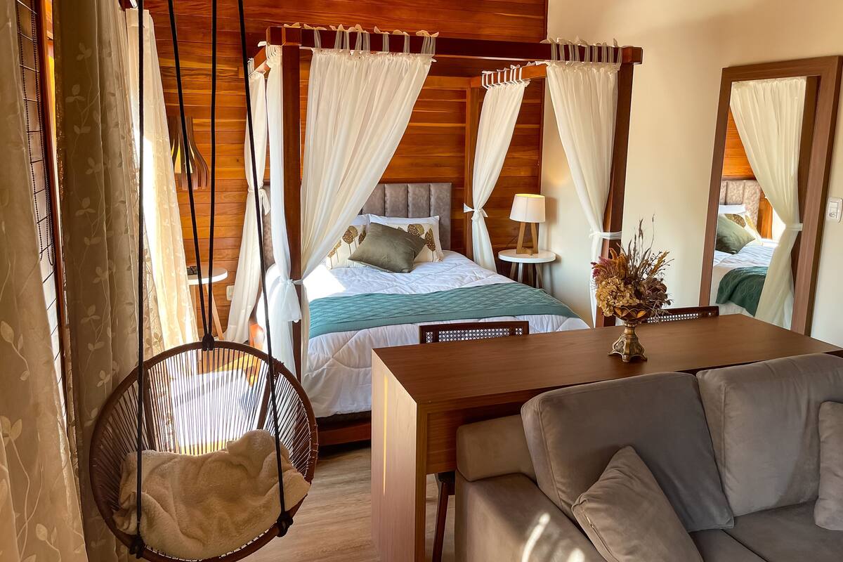 A room with a four-poster bed, a hanging chair, a sofa, and a small table with a lamp.