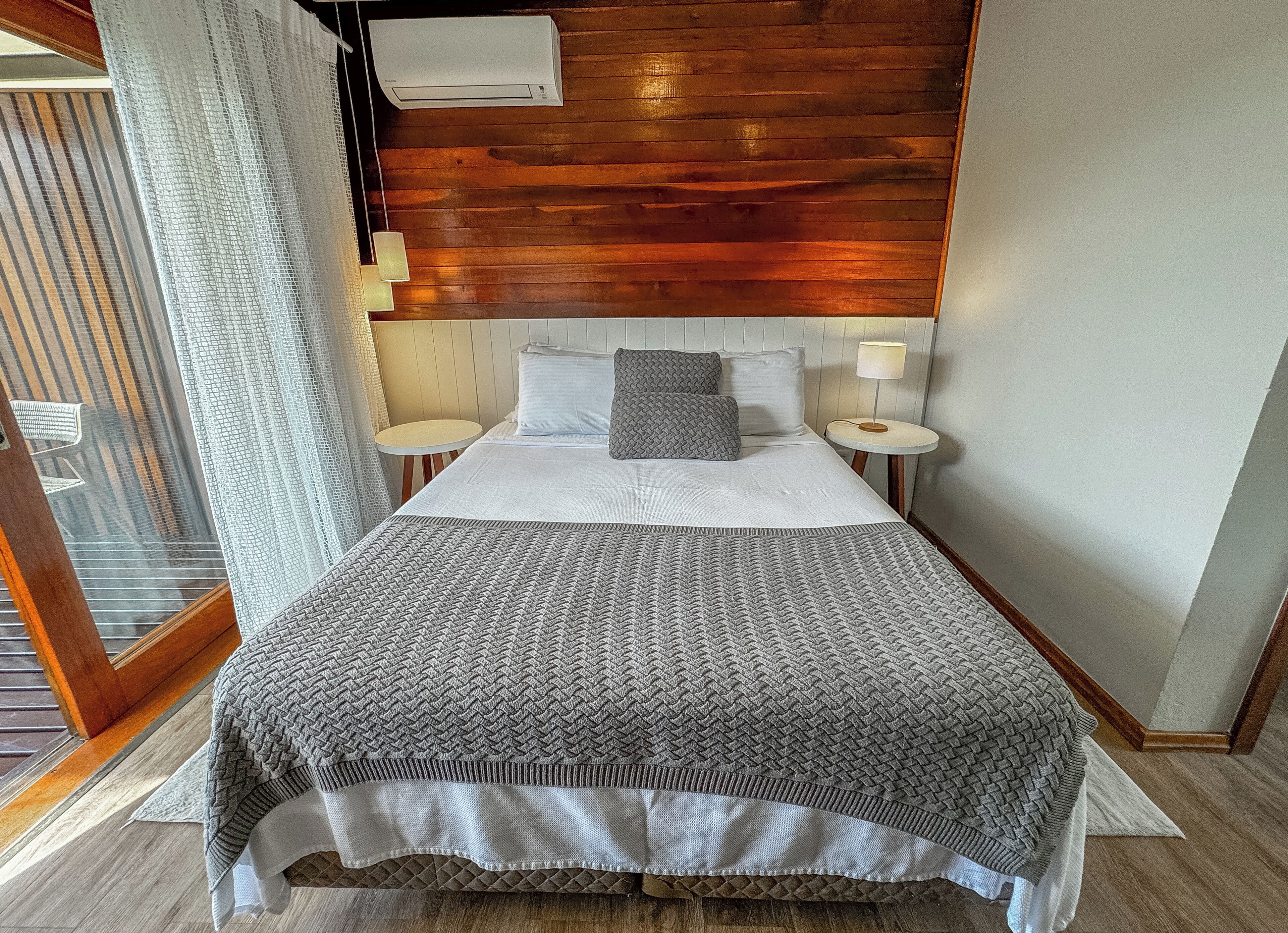 A hotel room with a large bed, a wooden headboard, two bedside tables with lamps, and a view through a window with curtains.