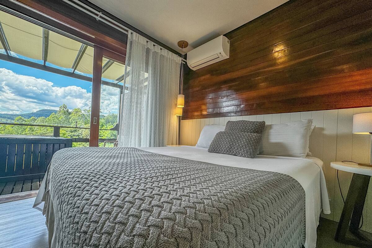 A bedroom with a large bed, a wooden headboard, a window with curtains, and a view of greenery.