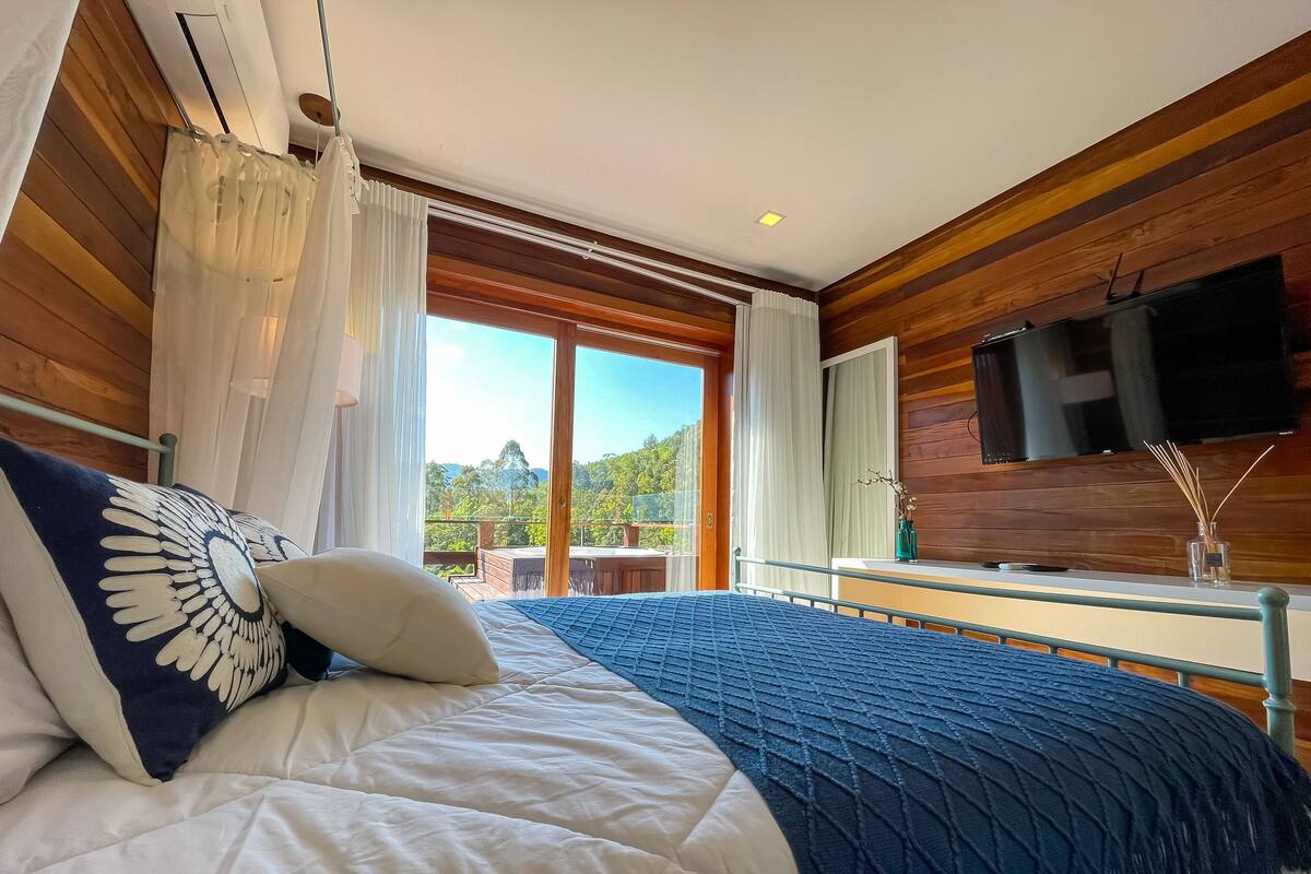 A bedroom with a large bed, wooden walls, a TV, and a balcony with a view of greenery.