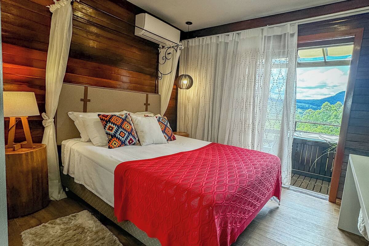 A bedroom with a bed, a nightstand, a lamp, a window with curtains, and a view of the outdoors.