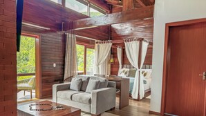 Ein gemütliches Zimmer mit Holzbalkendecke, einem Bett mit weißen Vorhängen, einem grauen Sofa und einem Holztisch.