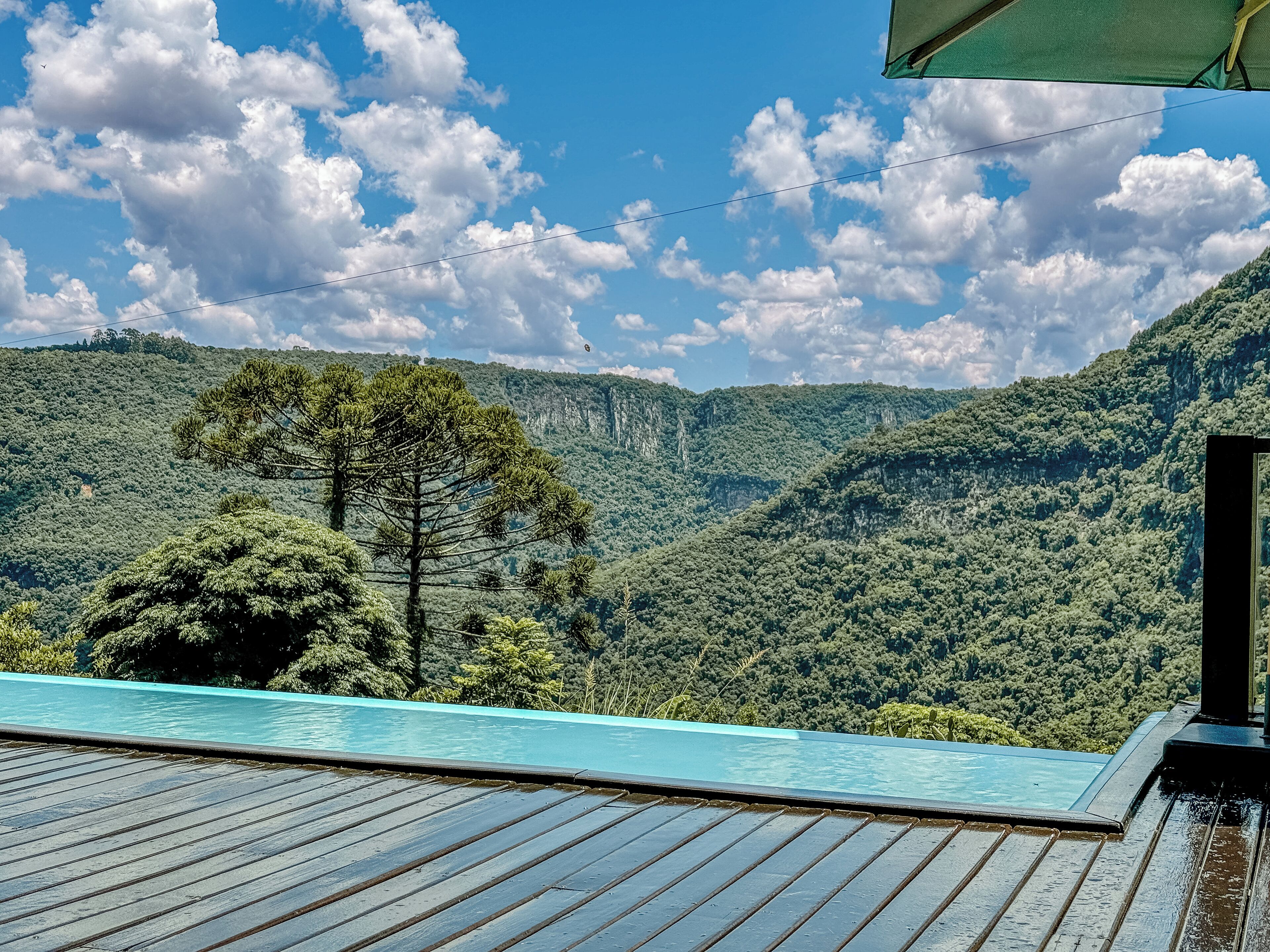 Vista a la piscina con terraza de madera, que domina un exuberante valle verde y una cadena montañosa bajo un cielo parcialmente nublado.
