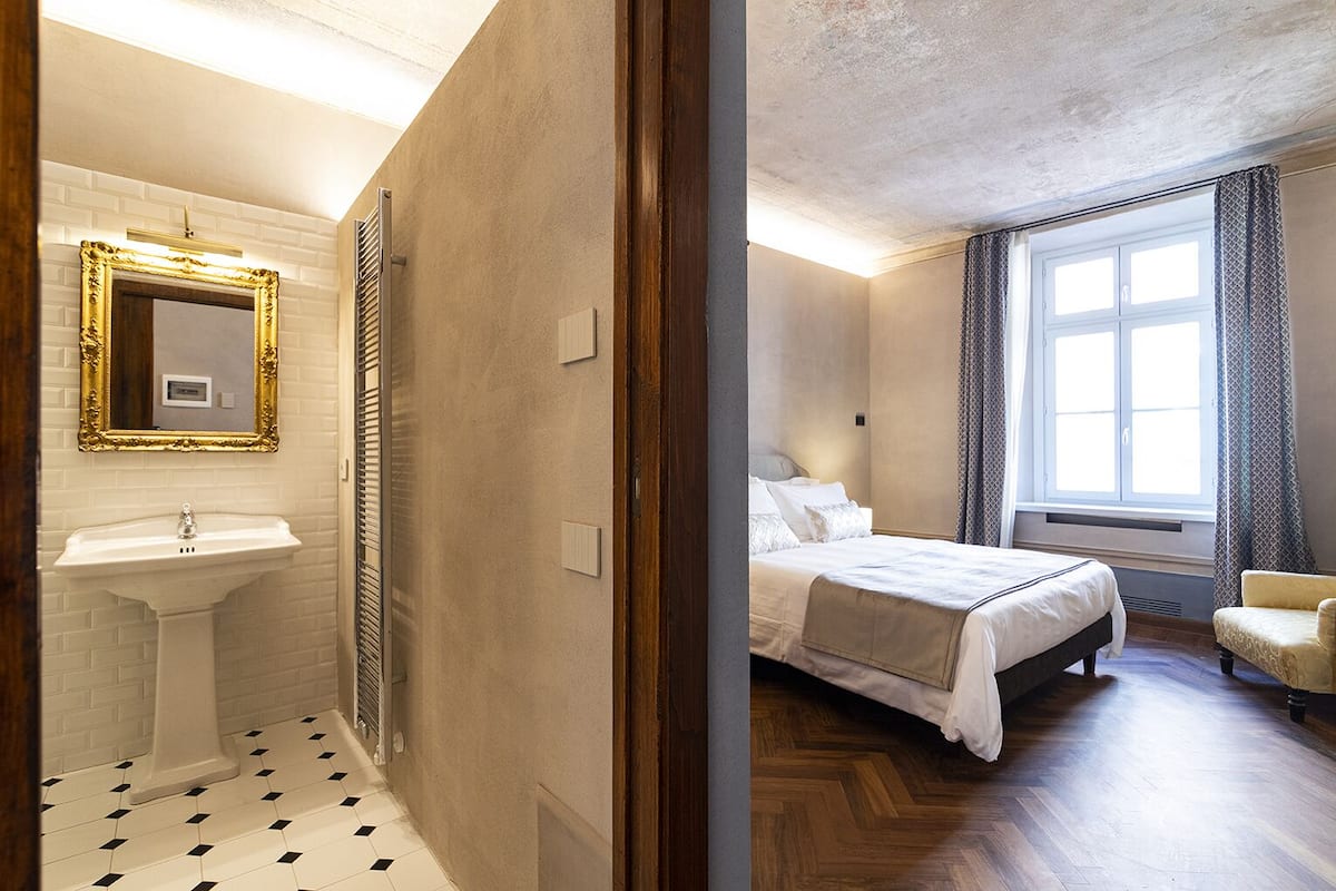 A hotel room with a bathroom featuring a sink, mirror, and radiator. The room has a bed, a window with curtains, and a chair.