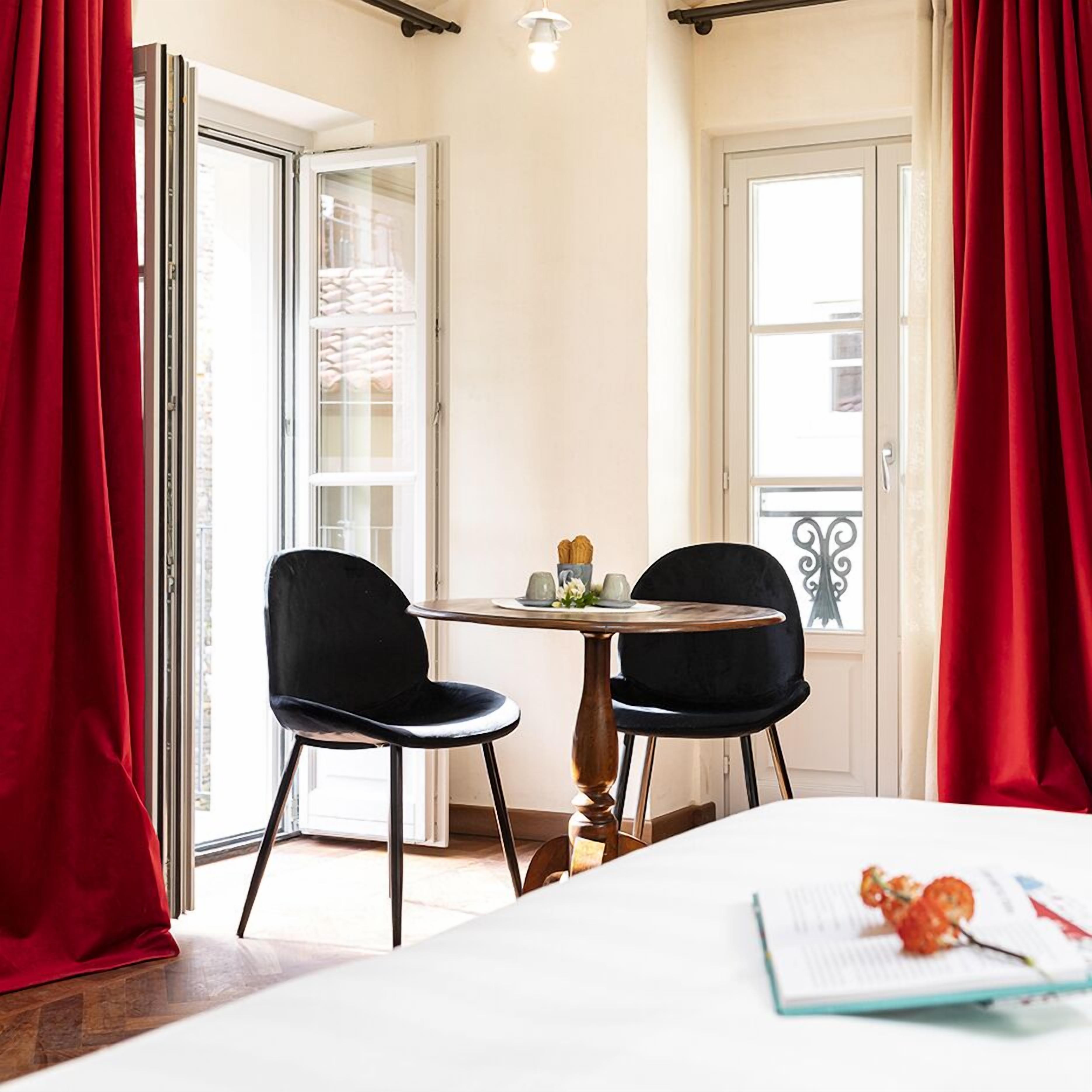 Habitación de hotel con una cama, dos sillas negras, una mesita con un florero y flores, y cortinas rojas.
