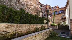 A stone retaining wall with a water feature, lush greenery, and traditional buildings with balconies in the background.