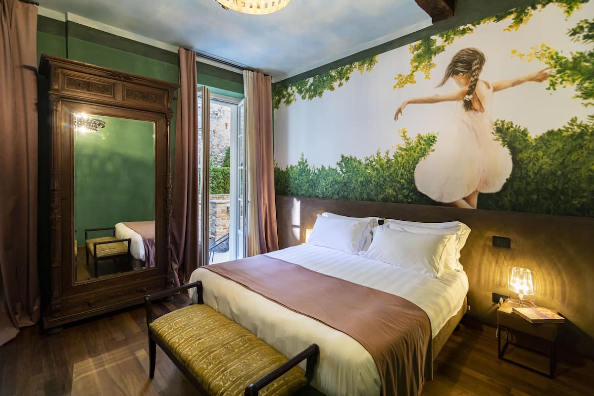 A bedroom with a large bed, a wooden headboard, a nightstand with a lamp, a mirror, and a view of greenery through a window.
