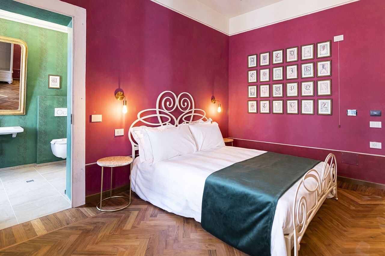A bedroom with a bed, a stool, a wall with framed pictures, and a mirror.