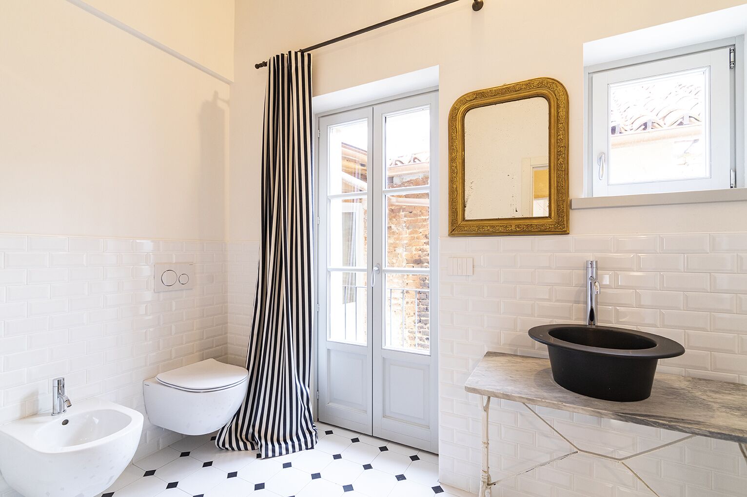 A bathroom with a white toilet, a bidet, a black sink with a black basin, a black and white striped shower curtain, and a gold-framed mirror above the sink.