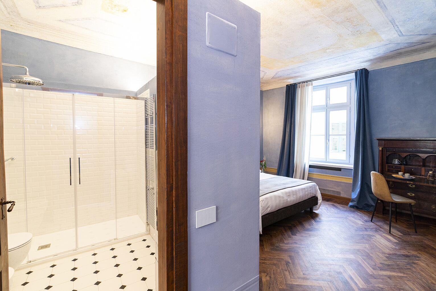 A bedroom with a bed, a dresser, a chair, a window with curtains, and a shower area with a tiled floor.