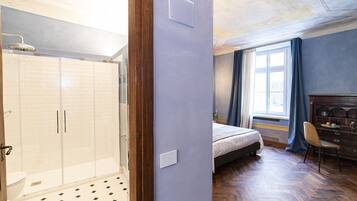 A bedroom with a bed, a dresser, a chair, a window with curtains, and a shower area with a tiled floor.