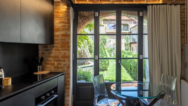 A modern kitchen with a glass dining table and chairs, a brick wall, and a view of a garden through a large window.