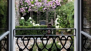 A view through a window with a wrought iron balcony railing, a brick building, and a garden with pink and white flowers.