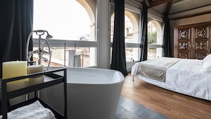 A modern bedroom with a large bathtub, a bed, and a view of the city through arched windows.