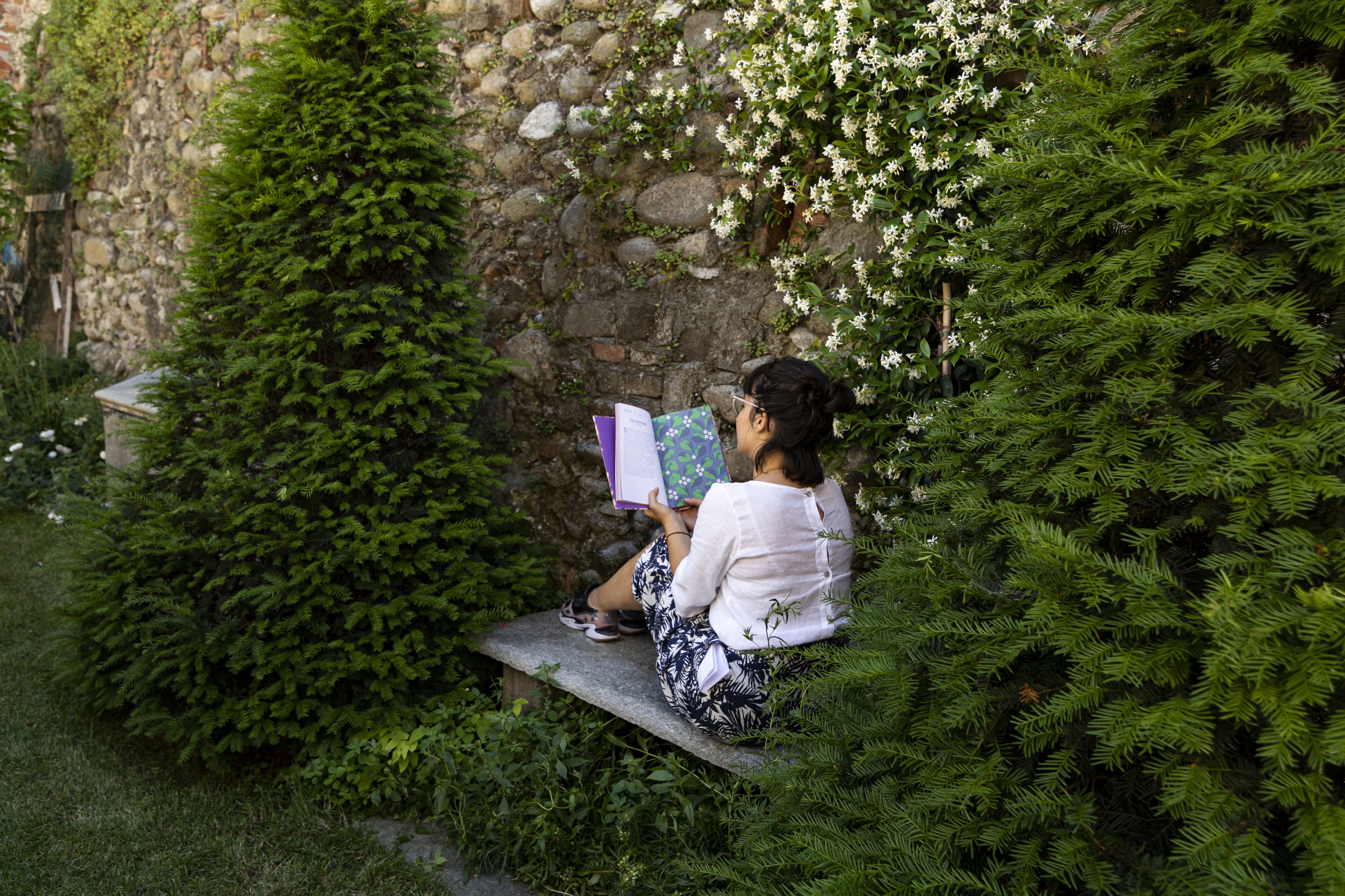 Uma pessoa sentada em um banco num jardim, com um muro de pedra e vegetação.