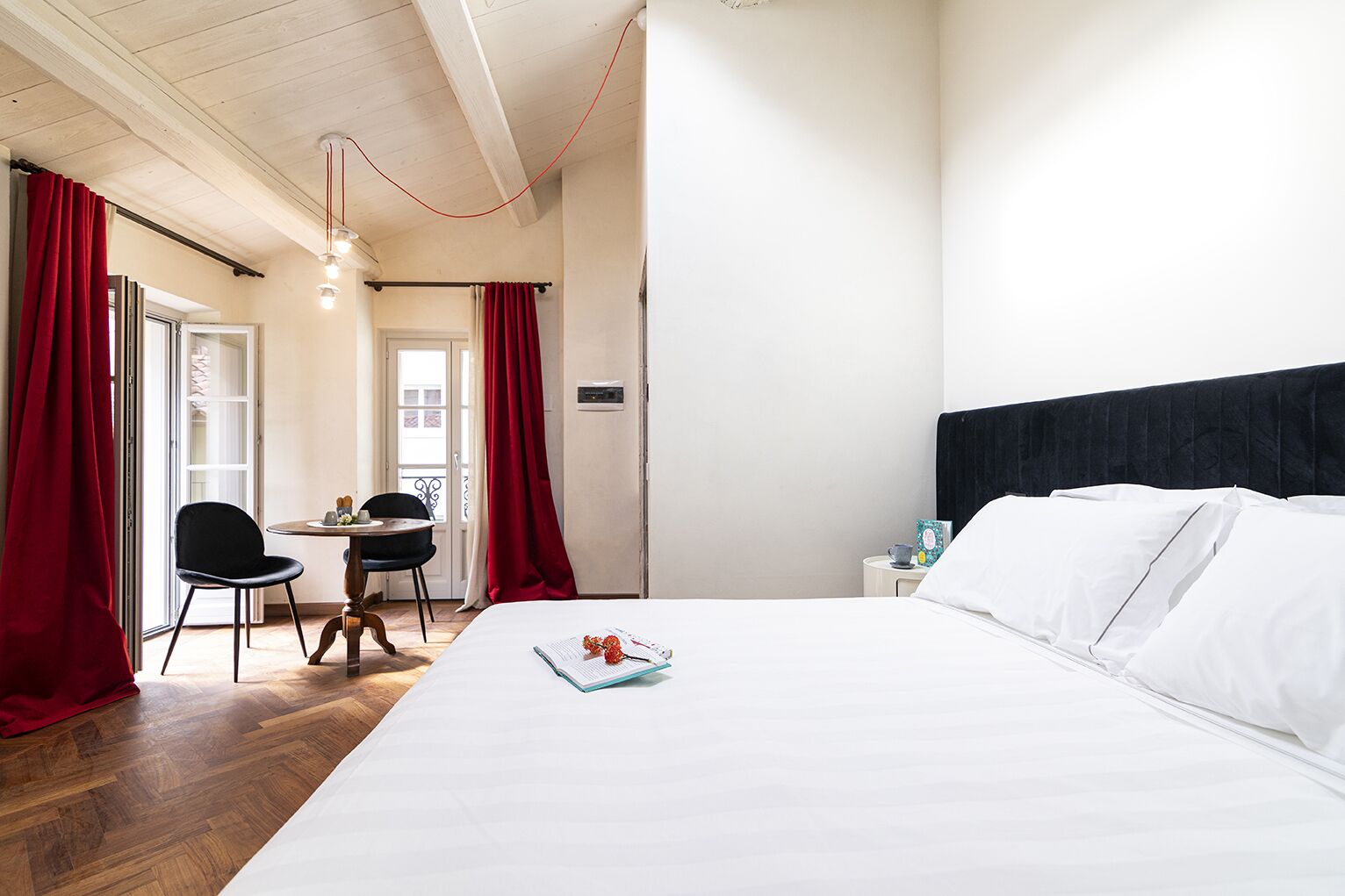 A hotel room with a bed, a desk with a chair, and a window with red curtains.