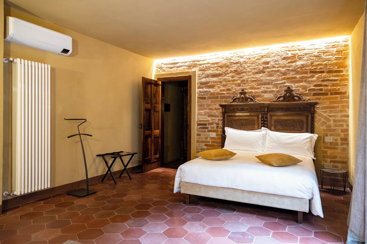 A bedroom with a brick wall, a bed with white bedding, a radiator, and a standing lamp.
