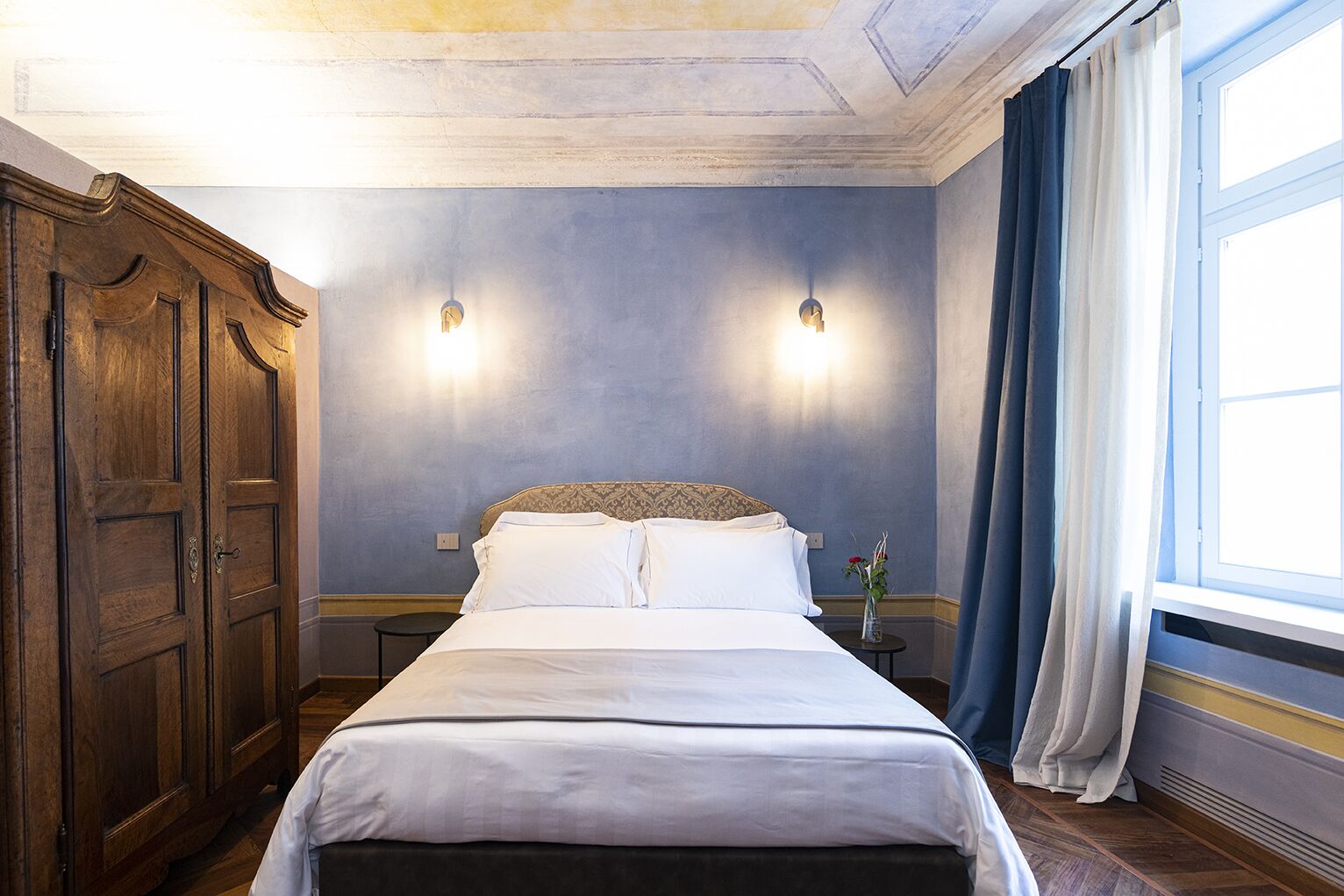 A hotel room with a large bed, wooden wardrobe, two wall sconces, a small table with a vase, and a window with curtains.