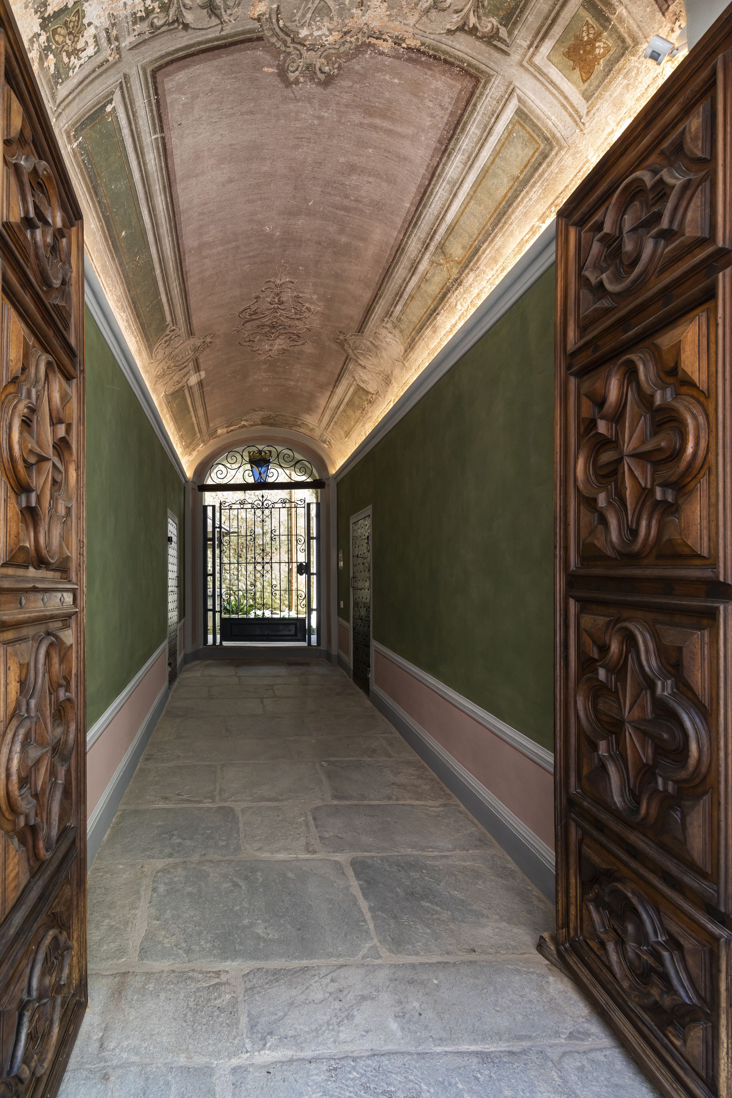 Un corridoio con porte in legno decorate e pavimento in pietra, che conduce a un ingresso chiuso da un cancello.