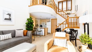 Ein geräumiges Wohnzimmer mit grauem Sofa, beigem Sessel und einem Esstisch mit Stühlen. Eine Holztreppe verbindet den Wohnbereich mit einer oberen Ebene.